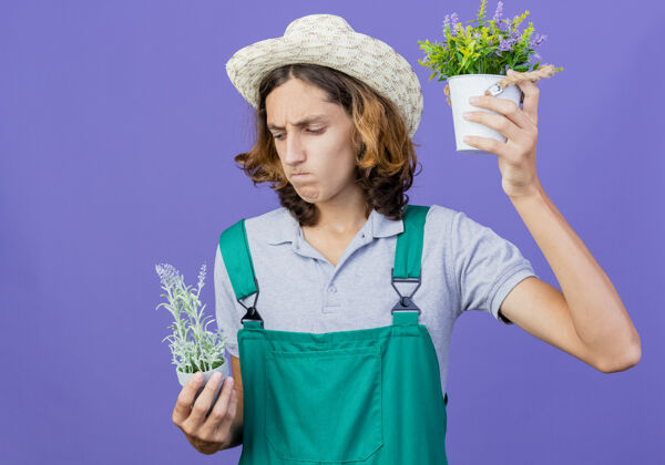 困惑年轻的园丁 穿着连体衣 戴着帽子 手里拿着盆栽植物花园连身衣穿着