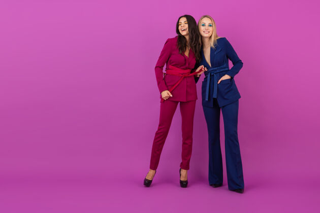 华丽高级时尚风格两位微笑迷人的女士在紫罗兰色的墙壁上穿着时尚多彩的紫色和蓝色晚礼服 朋友们一起玩 时尚潮流时尚年轻优雅