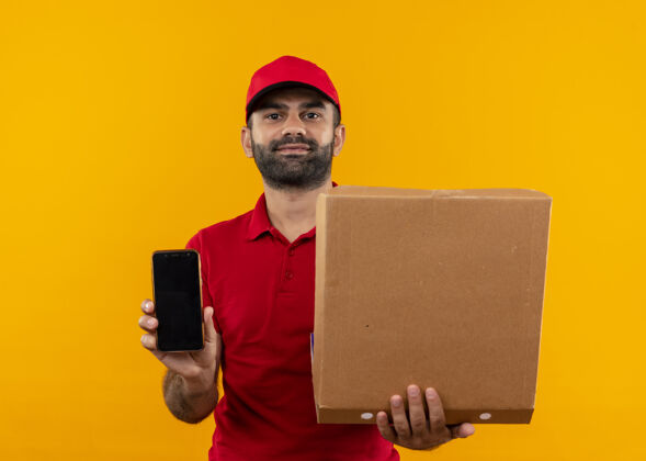 移动留着胡须的送货员身穿红色制服 戴着帽子 手里拿着打开的披萨盒 展示着站在橙色墙上自信表情的智能手机帽子制服胡须