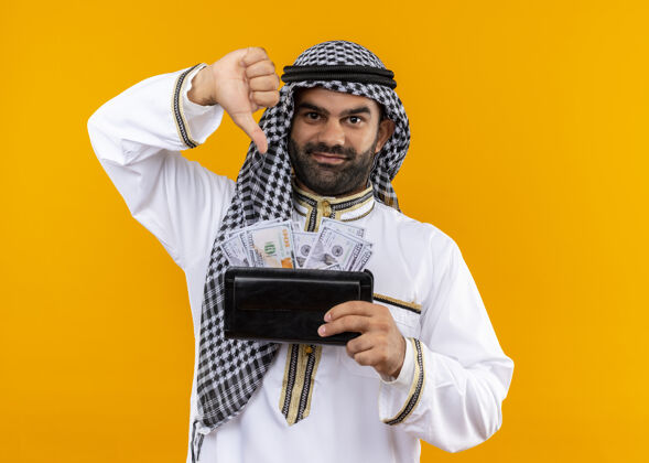 向下身着传统服饰的阿拉伯商人拿着钱包 面带微笑 自信地伸出大拇指站在橙色的墙上自信微笑商人