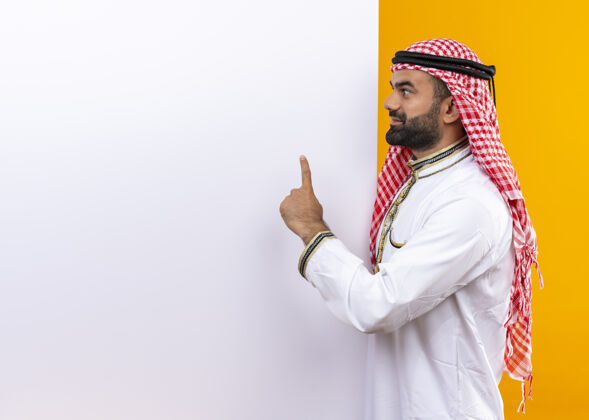佩戴身着传统服装的阿拉伯商人站在橙色的墙上 手举白色空白广告牌 面带微笑空白商务人士传统