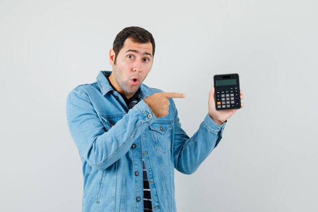 肖像一个穿着t恤衫和夹克衫的年轻人用手指着计算器 看上去很惊讶正视图成人时尚计算器