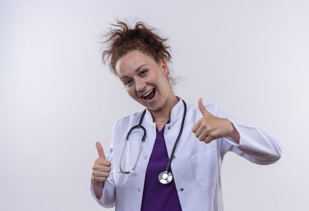医生身穿白大褂 手持听诊器的年轻女医生微笑着 双手竖起大拇指站在白色的墙上两者站立年轻人