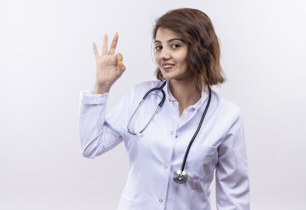 听诊器身穿白大褂 手持听诊器的年轻女医生微笑着 自信地站在白墙上做着“没事”的手势外套女人好的