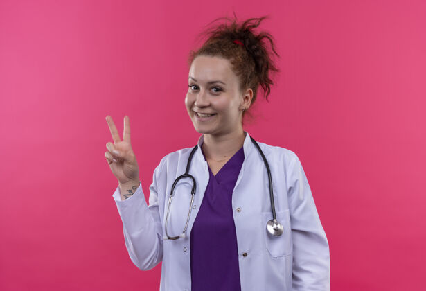 听诊器身穿白大褂 手持听诊器的年轻女医生微笑着 在粉红色的墙上立着胜利的牌子医生年轻外套
