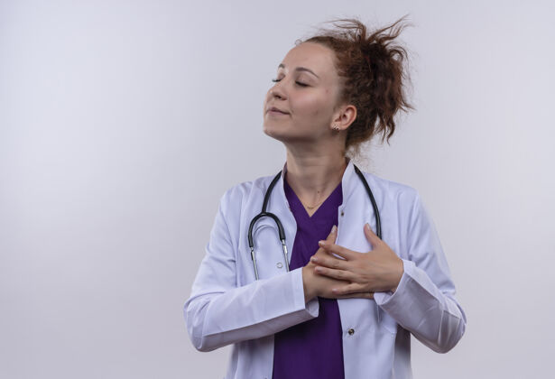 穿年轻女医生身穿白色外套 手持听诊器 双臂放在胸前 闭着眼睛站在白色的墙上 感受积极的情绪站着医生女人