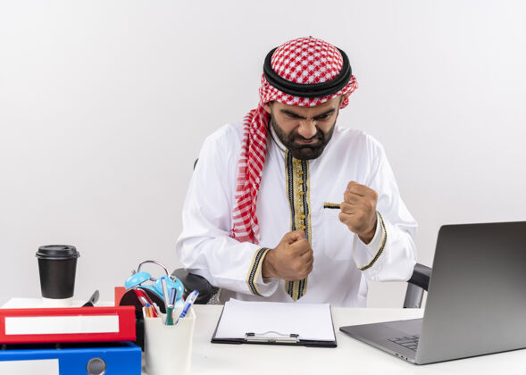 电脑身着传统服装的阿拉伯商人坐在办公室的桌子旁 用手提电脑握紧拳头 表情咄咄逼人 既不高兴又沮丧好斗商务人士笔记本电脑