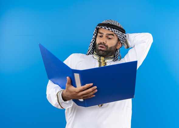 混乱阿拉伯商人在传统的穿着打开文件夹看着它困惑地站在蓝色的墙上商务人士站开放