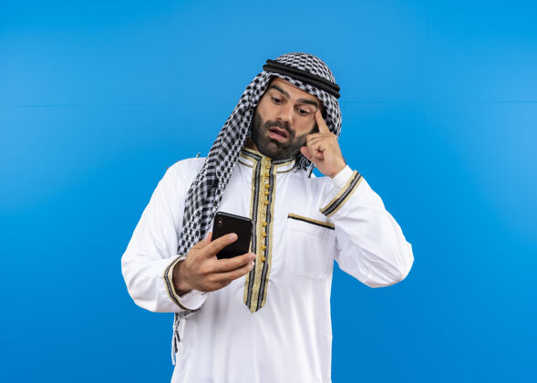 困惑一个穿着传统服装的阿拉伯人站在蓝色的墙上 看着自己手机的屏幕 困惑不已阿拉伯语穿移动