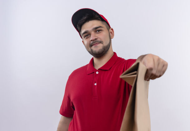 年轻年轻的送货员穿着红色制服 戴着帽子 手里拿着纸包 友好地微笑着包装制服拿着
