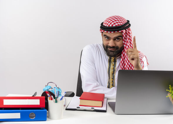 工作身着传统服装的阿拉伯商人坐在桌旁 拿着笔记本电脑 露出食指微笑 自信地在办公室工作阿拉伯语笔记本电脑坐着