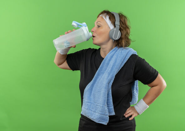 头带中年运动女性 身穿黑色t恤 头上系着头带 脖子上系着毛巾 锻炼后喝着绿墙上的水看起来很累年龄看中