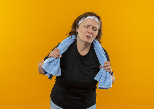 女人中年运动女性 身穿黑色t恤 头上系着头带 肩上搭着毛巾 站在橙色的墙上锻炼后 看上去既疲惫又疲惫头带肩膀疲倦