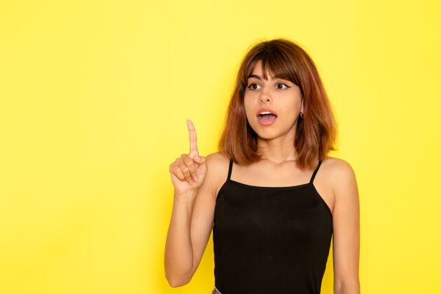 手指身穿黑色衬衫的年轻女性在浅黄色墙壁上抬起手指的正面视图姿势模特年轻