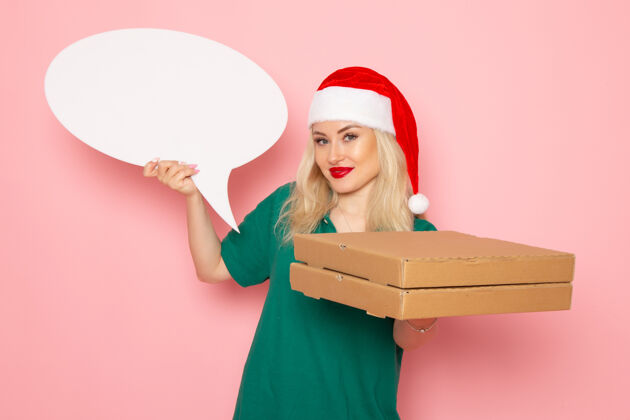 制服正面图年轻女性手持白色大牌子和食品盒在粉色墙上拍照工作制服新年假期工作快递圣诞节盒子视图