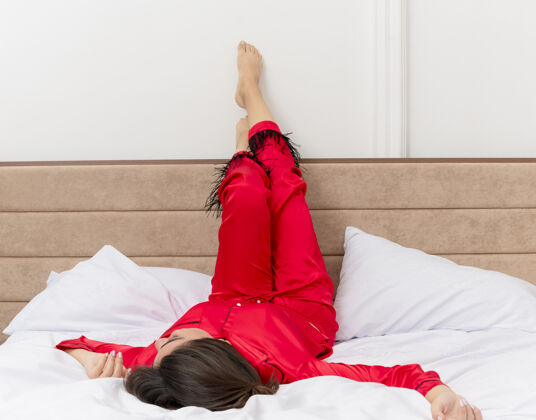 养穿着红色睡衣的年轻漂亮女人躺在床上 躺在柔软的枕头上 双腿翘起 愉快而积极地享受周末在卧室室内的灯光背景下红色享受快乐