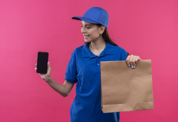 帽子穿着蓝色制服 戴着帽子 拿着纸包和智能手机的年轻送货女孩面带微笑地看着它递送纸年轻