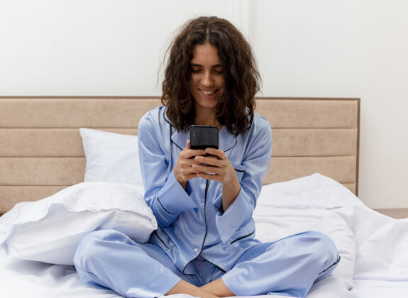睡衣穿着蓝色睡衣的有趣年轻漂亮女人坐在床上 用智能手机看着屏幕 在卧室内部的灯光背景下微笑着微笑移动屏幕