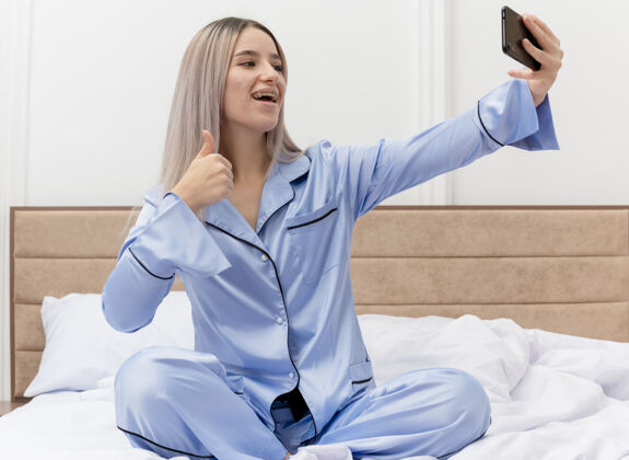 蓝色穿着蓝色睡衣的年轻美女坐在床上用智能手机自拍 在卧室内部灯光背景下竖起大拇指秀卧室室内