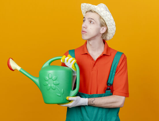 男人年轻的园丁穿着连体衣戴着帽子拿着浇水罐脸帽子园丁