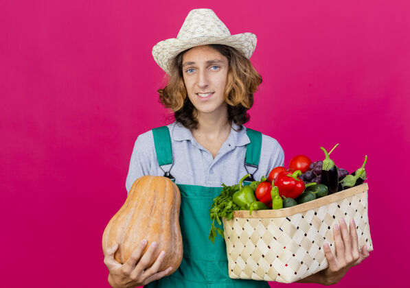 微笑年轻的园丁穿着连体衣戴着帽子拿着装满新鲜蔬菜的箱子抱着脸帽子