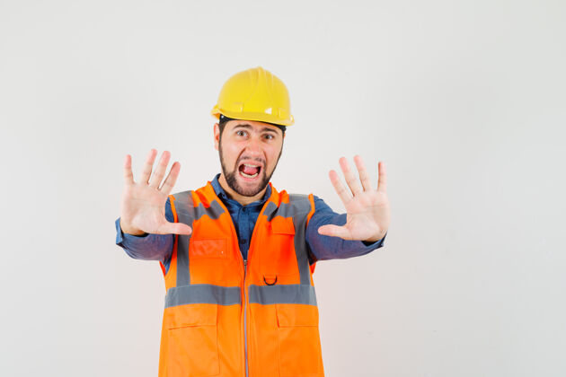 手势年轻的建筑工人穿着衬衫 背心 头盔显示停止手势 同时尖叫 看起来很害怕 前视图制服头盔衬衫