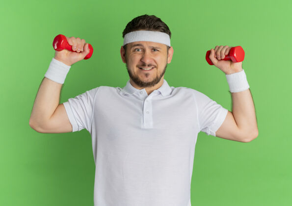 教练身穿白衬衫 头箍戴着哑铃的年轻健身男子站在绿色的墙壁上紧张而自信地锻炼身体紧张小伙子运动员