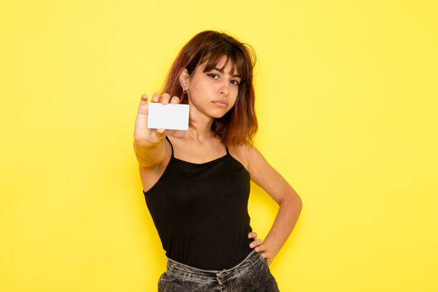 工作身穿黑色衬衫的年轻女性正拿着浅黄色墙上的白色塑料卡姿势情感女性