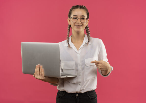 指着戴着眼镜 梳着辫子 穿着白衬衫的年轻女学生拿着笔记本电脑 手指指着笔记本电脑 微笑着自信地站在粉色的墙上人衬衫笔记本电脑