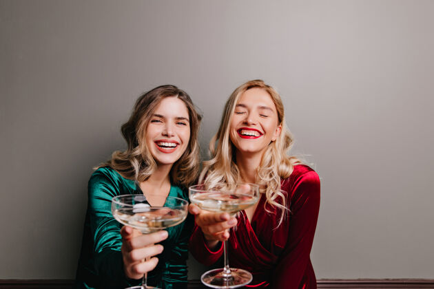 时尚穿着红色天鹅绒连衣裙的迷人白人女孩在喝香槟幸福的朋友在用酒庆祝年轻表情女人
