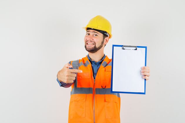 安全帽年轻的建设者穿着衬衫 背心 头盔指着剪贴板 看上去兴高采烈 俯瞰前方衬衫背心工作