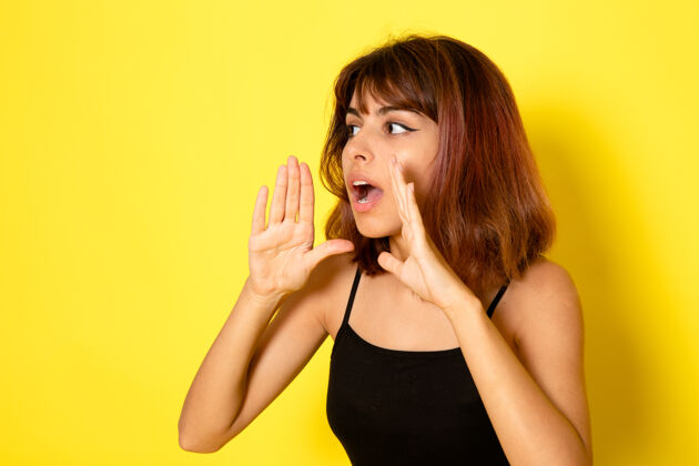 情感身穿黑色衬衫的年轻女性在浅黄色的墙上低声呼唤的正面视图女孩工作耳语