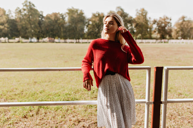 享受时尚的金发女郎在秋天的乡村享受着灿烂的阳光穿着红色套头衫和白色连衣裙的漂亮女孩在户外快乐地摆姿势明亮公园帽子