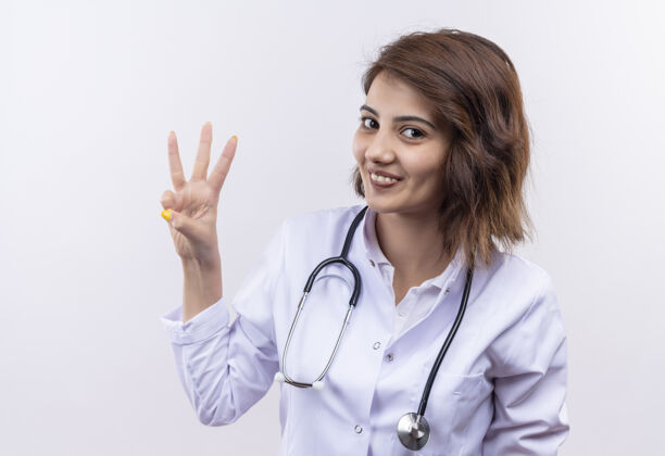 指指点点身穿白大褂 手持听诊器的年轻女医生站在白墙上微笑着指着三号手指手指外套站着