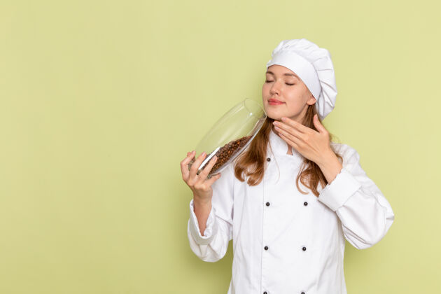 肖像身着白色厨师服的女厨师正拿着绿色墙上有棕色咖啡籽的罐子举行餐成人