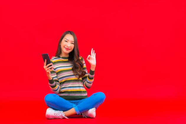 期待肖像美丽的亚洲年轻女子在红色隔离墙上使用智能手机科技销售信息