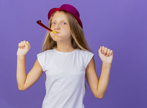 站着美丽的小女孩在节日帽子吹哨子握紧拳头高兴和兴奋 生日派对的概念站在紫色的背景兴奋帽子紫色