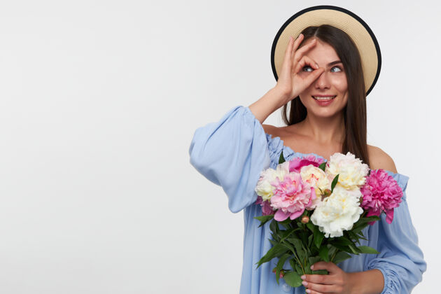 20多岁有吸引力的女孩的肖像 长黑发戴着帽子和蓝色的裙子手持一束美丽的鲜花 通过手指在白色墙上的复制空间左侧观看傻舌头花束