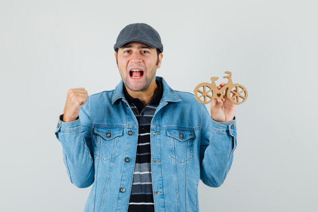 T恤一个穿着t恤 夹克 帽子的年轻人展示着赢家的姿态 手里拿着木制玩具自行车 看上去很幸运手势自行车木头