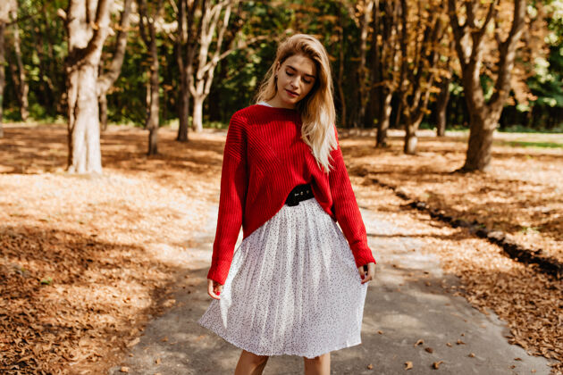 成人可爱的金发女孩在秋天的公园里摆姿势穿着漂亮的白裙子和漂亮的红毛衣叶子毛衣森林