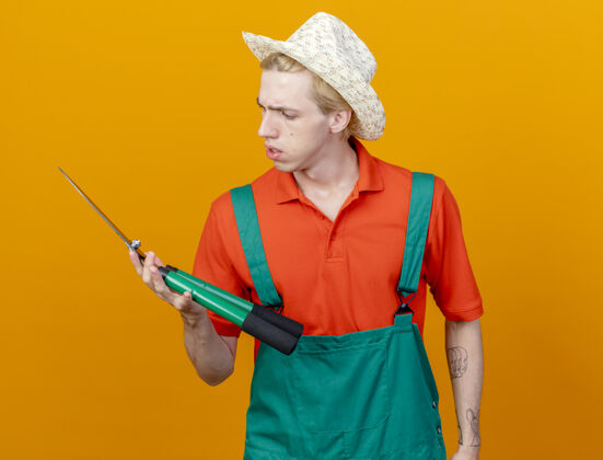 年轻年轻的园丁穿着连体衣戴着帽子拿着树篱剪看着剪着严肃的脸站在橙色的背景橙色男人严肃
