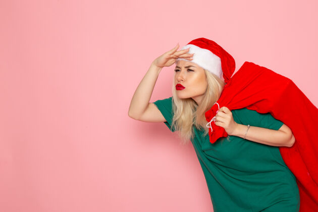 圣诞节正面图：年轻女性拎着红包在粉色墙壁上送礼物模型假日圣诞老人新年照片微笑美丽年轻女性