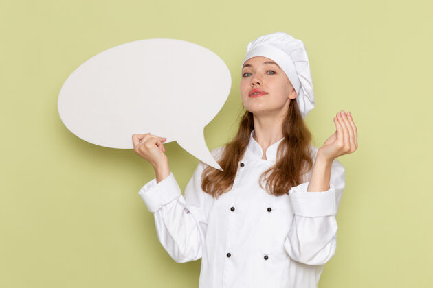 人女厨师身穿白色厨师服 手持绿色墙壁上的白色标志的正面视图举行肖像微笑