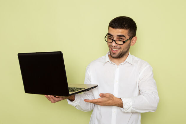 个人身穿白衬衫 手持笔记本电脑 在浅绿色墙壁上微笑的男性上班族的正视图手持微笑灯光