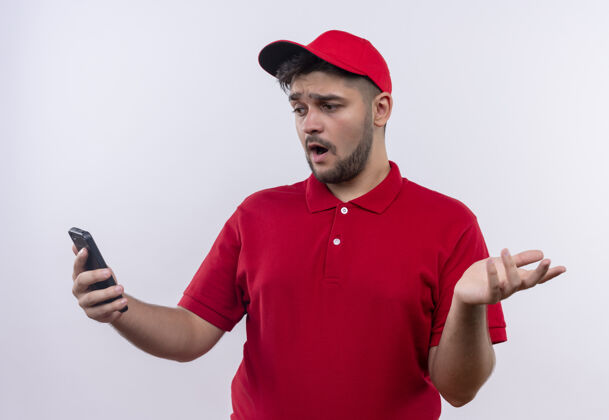 看年轻的送货员穿着红色制服 戴着帽子 看着智能手机的屏幕 显得困惑和不确定年轻男人站