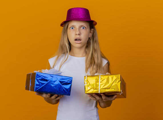 小漂亮的小女孩戴着节日礼帽拿着礼盒看着镜头惊讶又惊讶 生日派对的概念站在橙色的背景上漂亮生日站着