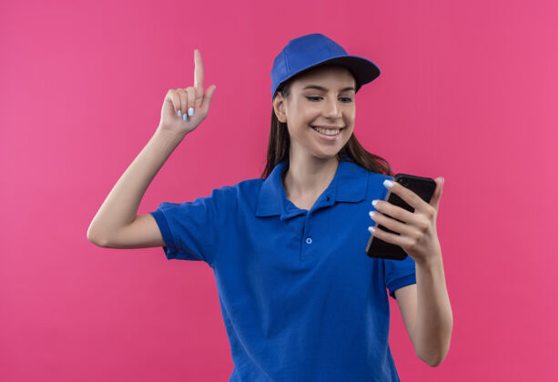 指向穿着蓝色制服 戴着帽子的年轻送货员看着手机屏幕 手指向上指着有着伟大想法的快乐帽子向上创意