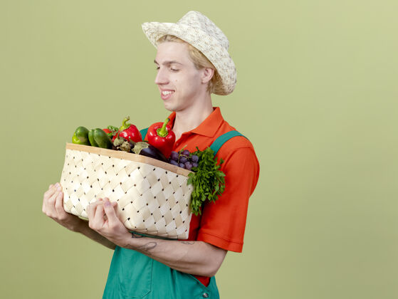 站着年轻的园丁 穿着连体衣 戴着帽子 手里拿着装满蔬菜的箱子 站在明亮的背景下 面带微笑地看着蔬菜满的连身衣脸