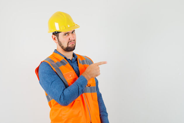 制服年轻的建设者指着边上穿着衬衫 背心 头盔 神情厌恶 前视工厂壁板勤杂工