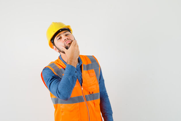 工程师年轻的建筑工人穿着衬衫 背心 头盔 摸着胡子 看着帅气的脸上的皮肤 前视皮肤帽子安全帽